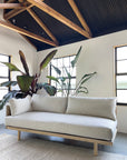 A modern, minimalist living room features the SOBU Woody Sofa (One Arm) in white ash with natural kapok cushions, set against large indoor plants, exposed wooden beams, black ceiling panels, white walls, and natural light from two windows.
