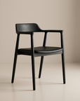 The SOBU Sven Dining Chair features a sleek black ash frame with a curved backrest, leather seat, armrests, and four straight legs. Styled against a plain beige background, this modern chair blends comfort and elegance.