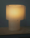 The Snegl Scallop Table Lamp by RUSSO BETAK STUDIO sits on a light wooden surface, its ribbed, biodegradable design casting a soft, elegant glow—bringing a story from the sea against a dark gray backdrop.