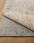 A corner of a patterned rug is lifted to reveal the Loloi Cushion Grip Rug Pad - 1/4, made from recycled synthetic fibers, both atop a light wood floor for enhanced durability.