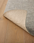 The Loloi Cushion Grip Rug Pad - 1/4, made from recycled synthetic fibers in gray with a tan textured underside, is shown partially lifted to display the light-colored wooden floor beneath.