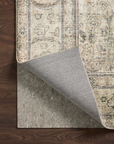 A beige patterned rug is partially lifted to reveal a gray Loloi Cushion Grip Rug Pad - 1/8, made from recycled synthetic fibers, underneath on a dark wood floor.