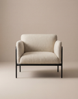 The Scribe Lounge Chair | Fabric by Wooden Stories features a contemporary design with beige upholstery, rounded armrests, and a sleek black metal frame, perfectly set against a neutral beige backdrop.