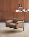 The Scribe Lounge Chair | Leather by Wooden Stories, featuring a modern beige design with a Bauhaus-inspired metal frame and black legs, sits on a textured rug before a wooden wall. A bar area behind displays bottles, glasses, and a small framed portrait.