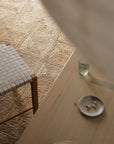 Minimalist scene with a woven chair on the Armadillo&Co Sequoia Rug in Natural, a light wooden table, an empty glass, and a small plate with a few nuts, partially obscured by a sheer white curtain.