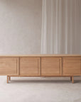 The Yorke Sideboard by Wooden Stories, crafted in solid oak with four paneled doors and slender legs, sits against a light beige wall with sheer white curtains, creating a simple and modern atmosphere.