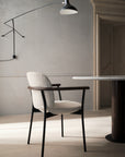 A modern, minimalist dining room featuring the Wooden Stories Solace Dining Armchair | Fabric, a round marble table, and a black wall lamp set against a neutral wall with a wooden door.