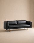 The Sonder Sofa | Leather by Wooden Stories—a modern black leather piece with Danish lines, slim proportions, and wooden legs—is set in an empty room with beige walls and floor.