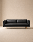 Centered against a plain beige background, the Sonder Sofa | Leather by Wooden Stories features modern Danish lines with a slim profile and wooden legs. Its black leather upholstery is complemented by feather-filled cushions, adding extra comfort to its contemporary design.