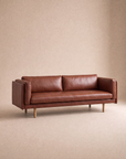 The Sonder Sofa | Leather by Wooden Stories, upholstered in modern brown leather with square arms and wooden legs, is set against a plain, light beige background. Its feather-filled cushions provide exceptional comfort while reflecting the timeless design sensibility of Borge Mogensen.