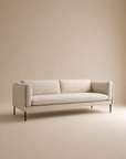 The Sonder Sofa | Fabric by Wooden Stories features modern Danish lines, a linear frame, thin black metal legs, and feather-filled cushions, elegantly set against a matching beige background.