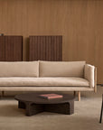 A minimalist living room with a Wooden Stories Sonder Sofa | Fabric with Leather Back in beige, featuring high-density foam cushions, a dark wood coffee table holding a book, two tall wooden cabinets, wooden wall paneling, and part of a dark armchair.
