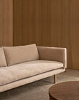 A Sonder Sofa | Fabric with Leather Back by Wooden Stories, featuring high-density foam cushions and wooden legs in a beige finish, sits on a gray rug beside a round wooden coffee table. Smooth wooden panel walls in the background create a warm, minimalist atmosphere.
