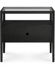 The Spindle Nightstand - Black Oak by Ethnicraft is crafted from solid European oak and features a glass top, an open shelf, a drawer with a round cut-out handle, and four slim legs for a modern look.