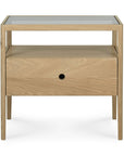 The Spindle Nightstand - Oak by Ethnicraft features solid oak construction, a glass top, an open shelf below, and a drawer with a central round cut-out handle.