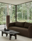 Modern living room featuring the Ethnicraft Figur Sofa | 3 Seater in brown, accompanied by a matching armchair, coffee table, and side table. Abstract wall art adorns the space, while expansive floor-to-ceiling windows reveal a lush forest view outside. Neutral tones and minimalist decor enhance the cozy, serene atmosphere—ideal for contemporary living.
