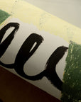 A close-up of THE POSTER CLUB’s Energize - Framed Print 20 x 28 shows bold black brushstroke lettering with green and yellow edges, displayed on a wooden surface.