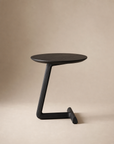 The SOBU Teena Side Table - Black Ash features a modern, minimalist design with a round black-stained ash top, single angled leg, and T-shaped base, shown in a compact form against a plain beige background.