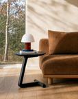 A modern living room features a brown suede sofa and the SOBU Teena Side Table in black ash, its cantilevered top displaying a small white and red lamp, books, and sunlight pouring through large windows with trees outside.