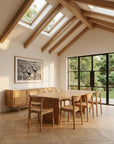 A modern dining room featuring the SOBU Tommaso Dining Table with wood legs, wooden chairs, large garden-facing windows, skylights, exposed beams, light wood flooring, and a sideboard topped by a black-and-white photo.