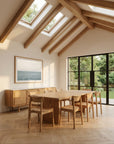 A bright dining room with a SOBU Tommaso Dining Table - Wood Legs, six matching chairs, a sideboard, large garden-facing windows, skylight with exposed beams, and a framed sea picture on the wall.