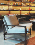 The Troubadour Lounge Chair | Fabric by Wooden Stories features light gray cushions, Danish lines, and a dark solid oak frame. Displayed in-store, it's surrounded by shelves of home decor, ceramics, and soft lighting.