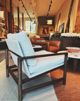A modern showroom displays the Wooden Stories Troubadour Lounge Chair | Fabric, featuring a light gray cushion and solid oak frame, surrounded by leather and fabric chairs, wooden tables, shelves, and warm ambient lighting.