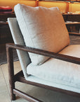 A close-up of the Troubadour Lounge Chair | Fabric by Wooden Stories, featuring light beige cushions and a dark wood frame, set indoors with shelves and folded textiles softly lit in the background.