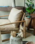 The Wooden Stories Troubadour Lounge Chair | Leather features a solid oak frame and leather cushions, positioned beside a wooden table and a potted plant in a cozy, sunlit room with neutral decor.