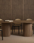 A modern dining room showcases the Wooden Stories Tathra Ellipse Dining Table with a stone top, accompanied by six black-framed chairs with woven cane backs, set against wood paneling and a beige carpet. An open book highlights European craftsmanship.