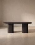 The Tathra Ellipse Dining Table by Wooden Stories, crafted from dark oak with two substantial cylindrical legs and an elegant oval silhouette inspired by classic Danish design, is showcased against a plain beige background.