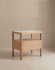 The Theo Nightstand Wide by Wooden Stories is a modern wood side table with Danish lines, a rectangular white top, single drawer, and four square legs, shown against a neutral beige textured wall and floor.