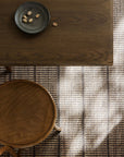 A top-down view of a wooden table with a dark plate holding a few nuts, one nut on the table, next to a wooden chair and Armadillo&Cos Trellis Rug in Natural & Bison with sunlight casting shadows.