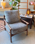 A stylish living space features the SOBU Viking Lounge Chair - Walnut with a gray textured cushion, placed next to a small table holding a lit lamp, books, and with a potted plant visible in the background.