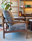 The Viking Lounge Chair - Walnut by SOBU, featuring plush textured gray cushions, sits beside a lamp-topped table. In the background, shelves with dishes and a potted plant highlight SOBU’s elegant furniture design.