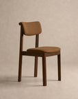 The Noki Dining Chair | Fabric from Wooden Stories is a contract grade piece crafted from solid wood, featuring brown fabric cushioning on the seat and backrest. Its simple, minimalist design includes straight legs and a gently curved back, shown here against a plain beige background.