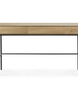 The Ethnicraft Whitebird Desk - Oak features a solid European oak top, two drawers, and slim black metal legs, displayed front-facing against a white background.