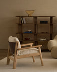 A minimalist living room featuring the Wooden Stories Wren Lounge Chair | Fabric in solid walnut, a beige armchair, a modern wooden bookshelf displaying books and bowls, and a gold decorative bowl atop it—all set against neutral walls and carpet, evoking timeless Danish furniture design.