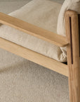 A close-up of the Wren Lounge Chair | Fabric by Wooden Stories, featuring light-colored upholstery and a beige textured carpet underneath. The chair showcases natural solid walnut arms and legs in a minimalist, sturdy design inspired by classic Danish furniture.