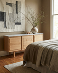 A minimalist bedroom featuring the Wooden Stories Yorke Sideboard with Stone Top, Scandinavian-style furniture, a vase of branches, textured wall art, a neutrally dressed bed, and natural light pouring through the window.