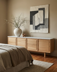 A minimalist bedroom features the Wooden Stories Yorke Sideboard with Stone Top, a large white vase with branches, abstract wall art, a bed with neutral linens, and decor in earthy tones for a simple Scandinavian-inspired look.