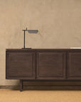 A minimalist room features the Yorke Sideboard by Wooden Stories, a black modern desk lamp, and a closed book atop it, all set against a beige textured wall and tan woven rug—perfectly capturing Scandinavian design.