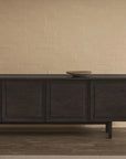 The Wooden Stories Yorke Sideboard in dark wood stands against a beige brick wall. A small brown lamp sits on the left and a shallow bowl on the right. Medium dark wood flooring enhances the Scandinavian aesthetic.