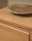 A close-up of the Yorke Sideboard by Wooden Stories, showcasing its light wood and simple Scandinavian design with a round beige ceramic plate on top against a neutral background with a textured wall.