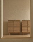 Two Yorke Highboards by Wooden Stories, featuring light wood and square panel designs, stand side by side against a beige brick wall in a softly lit, minimal room with wooden floors.