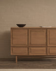 The Yorke Highboard by Wooden Stories stands against a beige brick wall, topped with a dark bowl and closed book, creating a simple, modern interior inspired by Danish design.