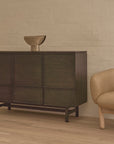 The Wooden Stories Yorke Highboard in dark wood with a minimalist sculpture on top stands beside a beige upholstered armchair in a room featuring light wooden floors and a white brick wall.