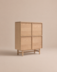 The Yorke Highboard by Wooden Stories features a light oak finish with four square doors and stands on a minimalist wooden frame. Isolated against a neutral beige background, this piece highlights its clean, modern aesthetic and visible wood grain, evoking classic Danish highboard style and Scandinavian design.