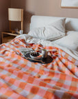 A cozy bed with white pillows features the FAIRE/TBCo Recycled Wool Blanket in Orange Gingham. On the bed, a tray holds a mug, cherries, and grapefruit beside a laptop. A wooden lamp sits on the bedside table.