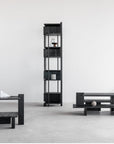 A minimalist room features Ethnicrafts Abstract Column dark wood bookshelf displaying a few books and objects, accompanied by matching dark wood benches and a coffee table on a light concrete floor, set against a plain white wall.